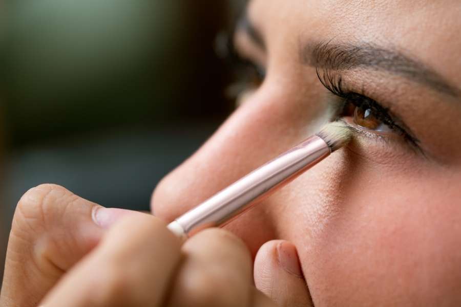 Gros plan sur une femme aux yeux bruns qui applique du fard au coin interne de l'œil avec un pinceau.
